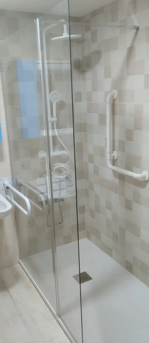 Ducha con mampara de vidrio, barra de apoyo blanca y asiento abatible